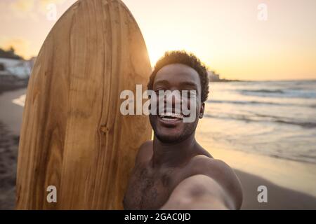 Happy afro surfer having fun taking selfie during sunset time - African man enjoying surf day - Extreme sport lifestyle people concept