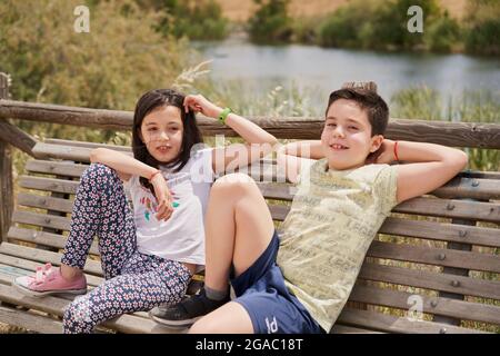 MOSTOLES, SPAIN - May 22, 2021: Children sitting on wooden bench laughing relaxed in park with pond in background Stock Photo