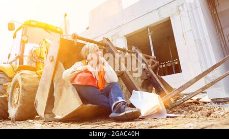 Female worker on construction site sits in shovel of excavator during her break when building a house Stock Photo
