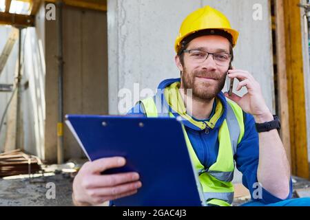 Craftsman on the construction site of building a house with a checklist while making a phone call with the smartphone Stock Photo