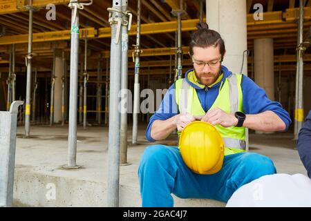 Young man as a construction worker with a yellow hard hat takes a break on the shell of a construction site Stock Photo