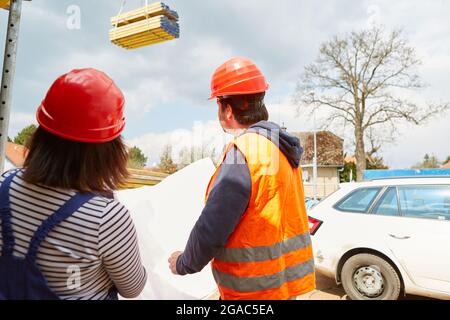 Worker and architect with architectural drawing watch crane unloading wood on construction site Stock Photo