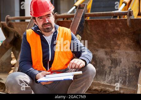 Craftsman or construction worker with checklist sitting on an excavator shovel at the construction site Stock Photo