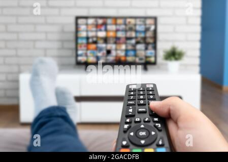 Man watching TV, lying on sofa, legs on table. Person holding remote control in living room Stock Photo