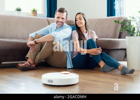 Robotic vacuum cleaner cleaning the room, smiling couple sitting on the floor. Smart home concept Stock Photo
