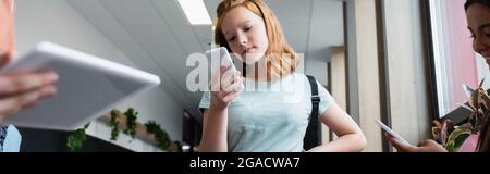 redhead girl using smartphone near blurred classmates with gadgets, banner Stock Photo