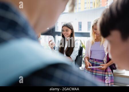 smiling schoolgirls using gadgets near boys on blurred foreground Stock Photo