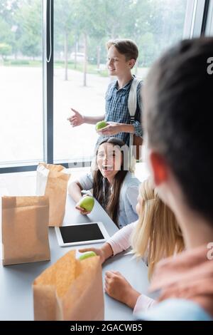 excited girl holding apple while laughing near classmates in eatery Stock Photo