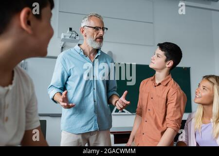 grey haired teacher gesturing while talking to teenagers in classroom Stock Photo