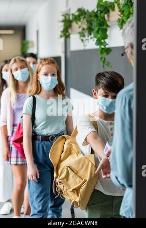 blurred teacher checking temperature of multiethnic pupils in medical masks with pyrometer Stock Photo
