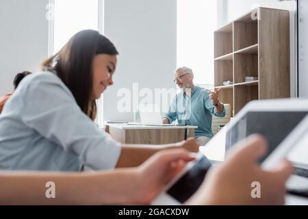 Smiling teacher using laptop and pointing with finger near blurred pupils Stock Photo
