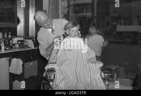 1940s, historical, a little boy sitting in a traditional barber's chair with a striped robe on getting a haircut, USA. Stock Photo