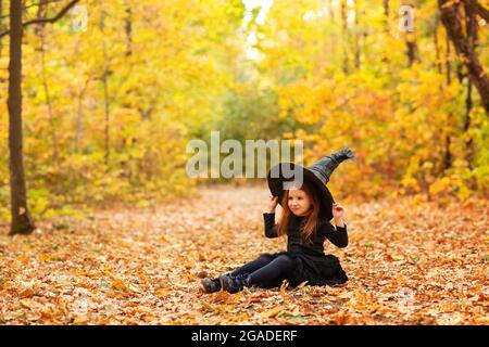 Cute happy little redhaired girl dressed in witch costume sitting on fallen leaves over autumn forest background with copy space, holding her hat with Stock Photo