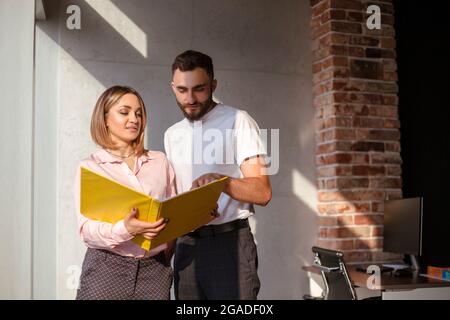 Two young focused coworkers, woman and man, looking through documents from folder standing in office. Business partners discussing financial graphs an Stock Photo