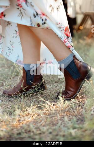 Cropped photo of female legs peeking out from under long, raised dress in mid-season natural leather dark brown flat ankle boots, dressed on grey sock Stock Photo