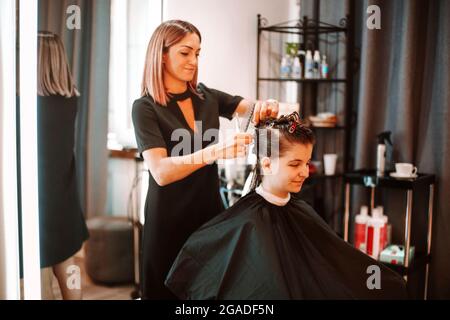 Positive woman hairdresser at working process in beauty salon, professional smiling hairstylist preparing female client for haircut, pinning up hair w Stock Photo