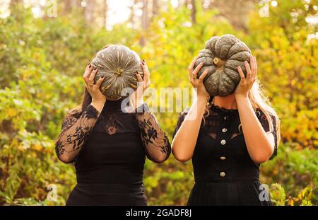 Two female friends holding scary green pumpkins in front of her face, standing isolated over autumn forest background. Fall holidays, halloween decora Stock Photo