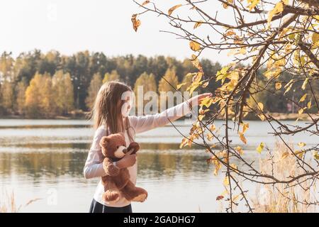 Pretty little girl iwith cute teddy bear holds dry leaf on riverbank in sunny forest on autumn day. Stock Photo