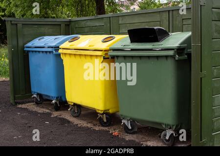 Three plastic multi-colored containers with rubbish near the fence. Separate collection of household waste. Responsible attitude to the environment. Stock Photo