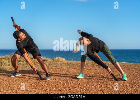 A middle-aged man and woman on the ocean are doing a warm-up. Fitness classes. Using gymnastic sticks for stretching. Stock Photo