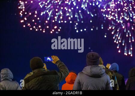 View from the back at unrecognizable people in warm clothes shooting fireworks on the phone at cold night. Stock Photo