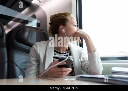 Thoughtful businesswoman listening to podcast on mobile phone while traveling by train. Stock Photo