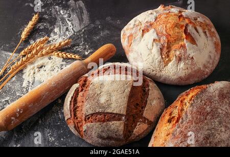 Wheat and rye bread loaves, flour, wheat ears and a wooden rolling pin on a black background Stock Photo