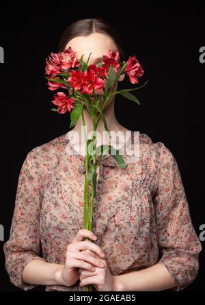Side view of young female in fred blouse hiding face behind natural flowers against black background Stock Photo