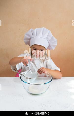 A little girl dressed as a cook pours flour from a glass into a glass bowl for kneading dough. Child development, child's lifestyle. The kid loves Stock Photo