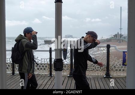 Brighton UK. 30 July 2021. Two teenagers battle against the wind and hold onto their hats on Brighton's Palace Pier. 50mph winds battered the south of England today. Large waves crashed onto the beach at Brighton. © Stuart Walden/ Alamy