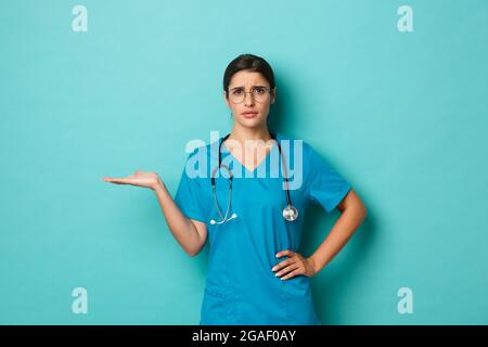 Coronavirus, pandemic and social distancing concept. Image of attractive female doctor in scrubs and glasses, raising hand and frowning confused, cant Stock Photo