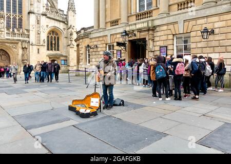 Bath, Somerset, UK - March 8 2016: A Busker playing a guitar and singing in front of Bath Abbey and the entrance to the Roman Baths Stock Photo