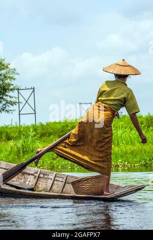 Burmese female wearing conical straw hat and colourful clothing leg rowing dugout canoe with one leg, Inle Lake, Myanmar Stock Photo