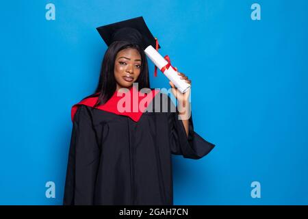 African-American beautiful woman in a robe and hat, on blue isolated background holding a diploma of higher education in her hands Stock Photo