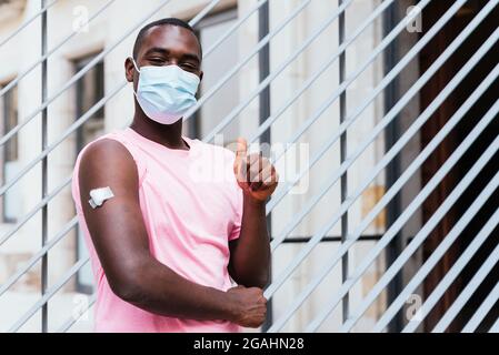 horizontal portrait of an african american young man. He is proud showing arm after vaccination and doing positive gesture with fingers Stock Photo