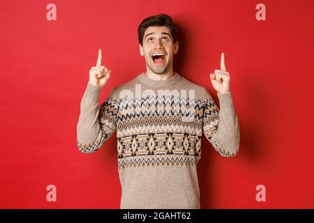 Image of excited handsome guy who likes winter holidays showing advertisement, looking and pointing fingers up with amazed face, standing in christmas Stock Photo