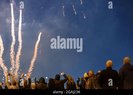 View from the back at unrecognizable people in warm clothes shooting fireworks on the phone at cold night. Stock Photo
