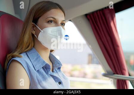 Travel safely on public transport. Close up of young woman with KN95 FFP2 protective face mask looking through bus window during her journey. Stock Photo