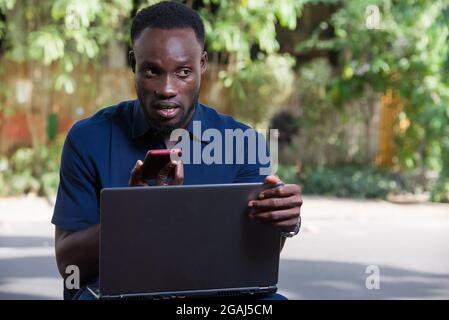 young man sitting outdoors with laptop computer communicating on cell phone while smiling. Stock Photo
