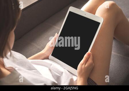 Girl sitting on sofa with digital tablet with black mockup screen, copy space Stock Photo