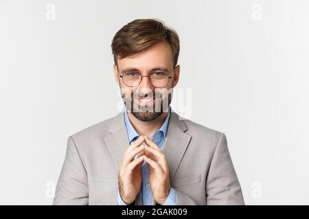 Close-up of bearded businessman in glasses and suit, scheming something, smiling devious and having perfect plan, standing over white background Stock Photo