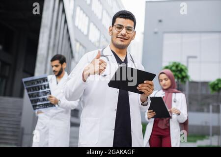Fornt view of smiling young arabic male doctor with tablet pc, standing outdoors near hospital building, showing thumb up. Two diverse colleagues standing behind him and working with xray Stock Photo