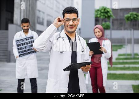 Coworking outdoors of hospital. Young handsome arab hindu man doctor in glasses using tablet device, posing to camera with smile. Multi ethnic male and female physicians working on the background Stock Photo