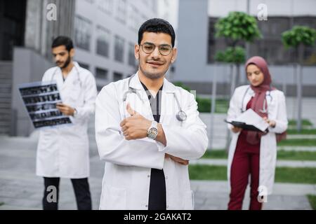 Fornt view of smiling young arabic male doctor, standing outdoors near hospital building, showing thumb up. Two diverse colleagues standing behind him and working with xray Stock Photo