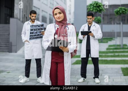 Female Muslim pleasant doctor in hijab, standing with clipboard outdoors in front of hospital building, while two handsome Arabian men doctors working with x-ray and tablet pc behind Stock Photo