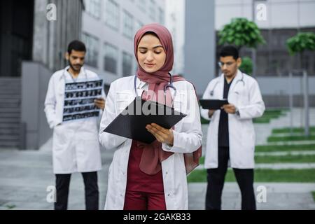 Pretty doctor in hijab writing notes on clipboard, standing ouside modern clinic. Two male Arab hindu doctors standing on the background, using digital tablet and examining patient's xray Stock Photo
