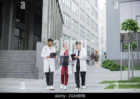 Three Arabian Muslim doctors, in medical suits and coats, discussing patient's x-ray tomography scan, walking outside on the background of modern hospital with stairs Stock Photo