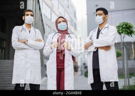 Front view of three multiethnic confident healthcare workers in medical suits and scrubs, wearing face masks, posing with arms crossed outdoors in front of modern hospital building. Covid-19 pandemy Stock Photo