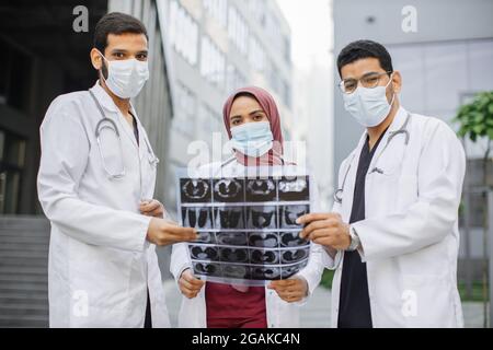 Diverse team of young medical scientists doctors neurologists, wearing uniform and mask, posing outdoors near hospital and looking at CT, MRI Scan. Stock Photo