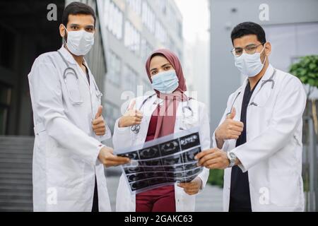 Team of three young diverse experts doctors in uniforms scrubs and protective masks, holding MRI x-ray tomography picture in front of modern clinic outdoors, looking at camera with thumbs up Stock Photo